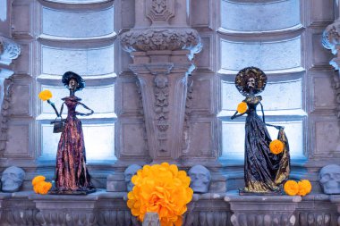 Two statues of women, each holding a flower. One of the statues is wearing a yellow dress. Day of the Dead celebration in Mexico, papel picado crafts, offerings, La Catrina