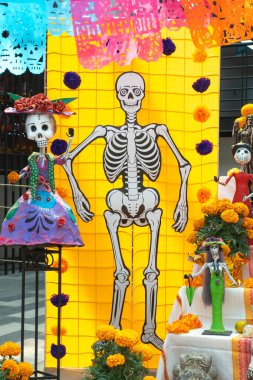 A skeleton is standing in front of a yellow wall with a skeleton on the wall behind it. Day of the Dead celebration in Mexico, papel picado crafts, offerings, La Catrina
