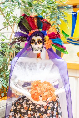 A skeleton is dressed in a purple veil and a flower garland, giving it a festive and colorful appearance. Day of the Dead celebration in Mexico, papel picado crafts, offerings, La Catrina