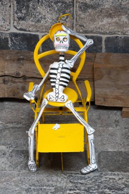 A skeleton is sitting on a yellow chair. The skeleton is holding a cup in its hand. Day of the Dead celebration in Mexico, papel picado crafts, offerings, La Catrina