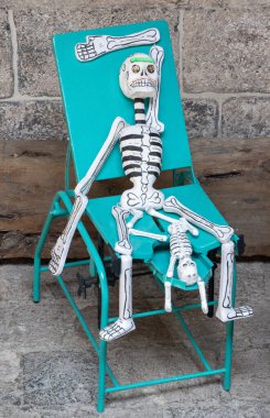 A skeleton is sitting on a green chair with another skeleton on top of it. The chair is made of metal. Day of the Dead celebration in Mexico, papel picado crafts, offerings, La Catrina
