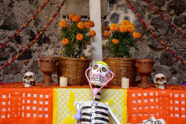 A skeleton is sitting on a table with a basket of flowers and skulls. The skeleton is wearing a stethoscope. Day of the Dead celebration in Mexico, papel picado crafts, offerings, La Catrina