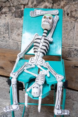 A skeleton is sitting on a chair with a skeleton on top of it. The skeleton on top is smaller and has a green face. Day of the Dead celebration in Mexico, papel picado crafts, offerings, La Catrina