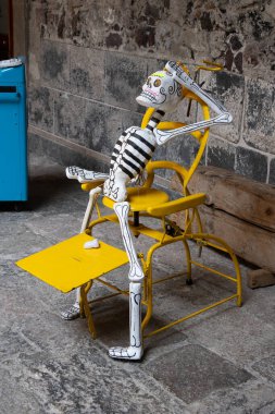 A skeleton is sitting on a yellow chair. The skeleton is wearing a black and white striped shirt. Day of the Dead celebration in Mexico, papel picado crafts, offerings, La Catrina