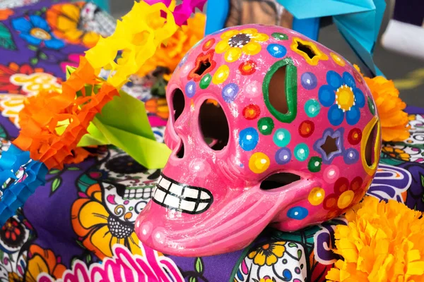 A pink skull with flowers painted on it sits on a table. The skull is surrounded by colorful paper flowers. Day of the Dead celebration in Mexico, papel picado crafts, offerings, La Catrina