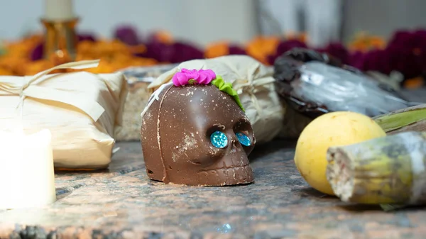 Bir çikolata kafatası diğer eşyaların olduğu bir masada. Kafatası mavi gözlerle süslenmiş ve kafasında bir çiçek var. Meksika 'da Ölüler Günü kutlaması, papel picado el sanatları, adaklar, La Catrina