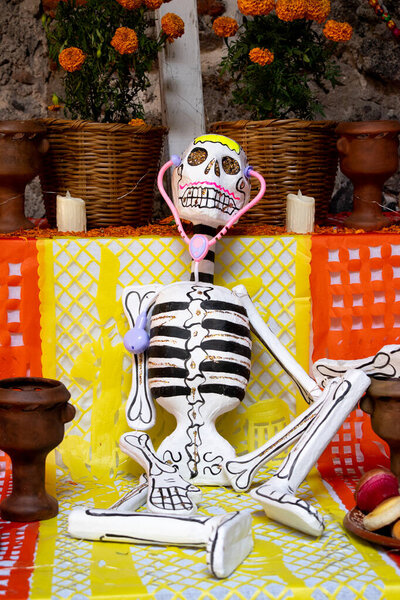 Скелет сидит на столе со свечами и цветами. Скелет украшен розовым и белым лицом. Празднование Дня Мертвых в Мексике, ремесла из папеля пикадо, подношения, La Catrina