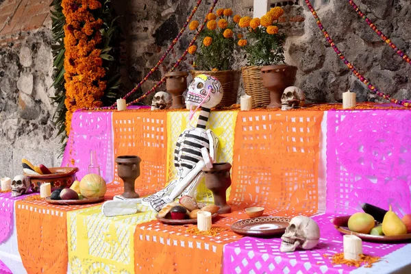 A table with a skeleton and skulls on it. The table is decorated with orange and pink tablecloths. Day of the Dead celebration in Mexico, papel picado crafts, offerings, La Catrina