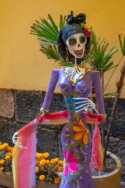 A woman dressed in a purple dress with flowers and a skeleton on her head. The skeleton is smiling. Day of the Dead celebration in Mexico, papel picado crafts, offerings, La Catrina