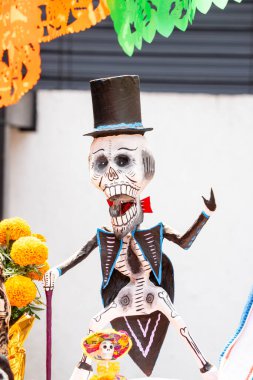 A skeleton dressed in a suit and top hat is holding a flower. The skeleton is smiling and he is happy. Day of the Dead celebration in Mexico, papel picado crafts, offerings, La Catrina