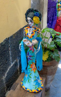 A skeleton dressed in a blue dress with flowers and a flower crown. The skeleton is holding a cup in one hand. Day of the Dead celebration in Mexico, papel picado crafts, offerings, La Catrina