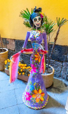 A woman in a purple dress with flowers painted on it. She is dressed in a skeleton costume. Day of the Dead celebration in Mexico, papel picado crafts, offerings, La Catrina