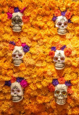 A wall of orange flowers with six skulls on it. The skulls are smiling and have purple and pink flowers on them. Day of the Dead celebration in Mexico, papel picado crafts, offerings, La Catrina