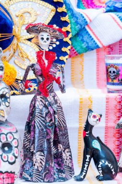 A woman dressed in a skeleton costume stands next to a cat. The cat is black and has a skeleton on its head. Day of the Dead celebration in Mexico, papel picado crafts, offerings, La Catrina