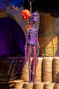 A purple and red skeleton is hanging from a string. The skeleton is made of paper and has a skull on its head. Day of the Dead celebration in Mexico, papel picado crafts, offerings, La Catrina