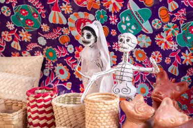 Two skeletons are standing next to each other on a purple background. The skeletons are dressed in white. Day of the Dead celebration in Mexico, papel picado crafts, offerings, La Catrina