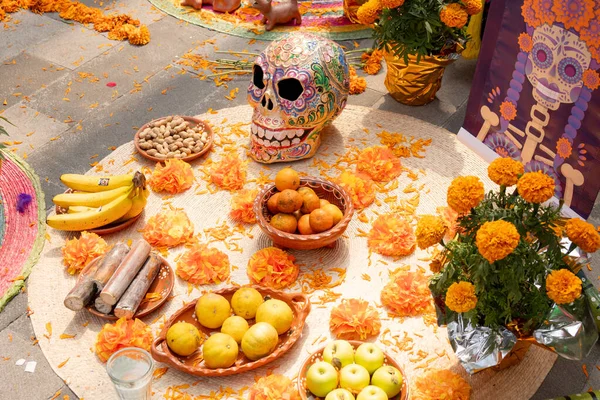 A table with a skull and fruit on it. The skull is decorated with flowers and the fruit is arranged in bowls. Day of the Dead celebration in Mexico, papel picado crafts, offerings, La Catrina