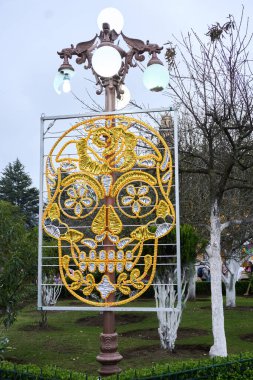 A large, ornate skull is suspended from a lamp post in a park. The skull is surrounded by a variety of flowers and leaves. Mexico, zacatlan de las manzanas, puebla, day of the dead mexican tradition