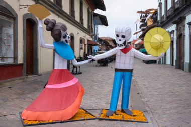 Two skeletons dressed in costumes are dancing in front of a building. The scene is set in a town square with a few chairs. Mexico, zacatlan de las manzanas, puebla, day of the dead mexican tradition