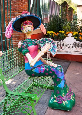A skeleton dressed in a green dress sits on a green bench. The skeleton is holding a fan and has a flower on its head. Mexico, zacatlan de las manzanas, puebla, day of the dead mexican tradition