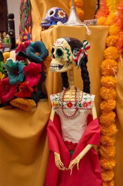 A skeleton dressed in a red dress and a white blouse is sitting on a table. The skeleton is surrounded by flowers. Mexico, zacatlan de las manzanas, puebla, day of the dead mexican tradition