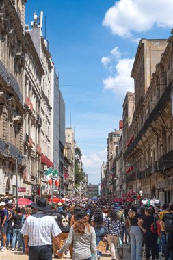 A busy street with a large crowd of people walking down it. The people are carrying bags and backpacks, and there are umbrellas and flags in the scene. Mexico City, Zocalo, historic building