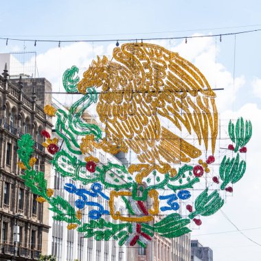 A large, colorful, ornate emblem with a bird on it hangs from a building. The bird is surrounded by leaves and flowers. Decoration to celebrate Independence Day in Mexico, zocalo