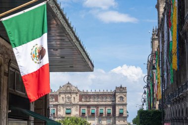 A flag with the colors red, white, and green is hanging from a building. The flag is in front of a building with a lot of windows. Decoration to celebrate Independence Day in Mexico, zocalo