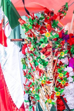 A colorful arrangement of flowers and leaves is displayed in front of a red and white flag. The arrangement is made of paper. Decoration to celebrate Independence Day in Mexico, zocalo