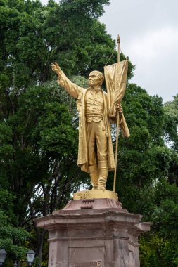 A statue of a man holding a flag. The statue is gold and is located in a park. Historic center of Queretaro City, decorations and traditions to celebrate Mexico's Independence Day, colonial architecture, alleys and facades