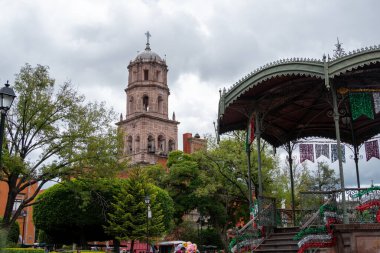 A large building with a tall tower and a green roof. The building is surrounded by trees. Historic center of Queretaro City, decorations and traditions to celebrate Mexico's Independence Day, colonial architecture, alleys and facades