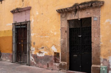 Two doors with bars on them are on a yellow building. The building is old. Historic center of Queretaro City, decorations and traditions to celebrate Mexico's Independence Day, colonial architecture, alleys and facades
