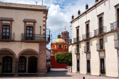 A small town square with a red building in the middle. The building has a dome on top. Historic center of Queretaro City, decorations and traditions to celebrate Mexico's Independence Day, colonial architecture, alleys and facades