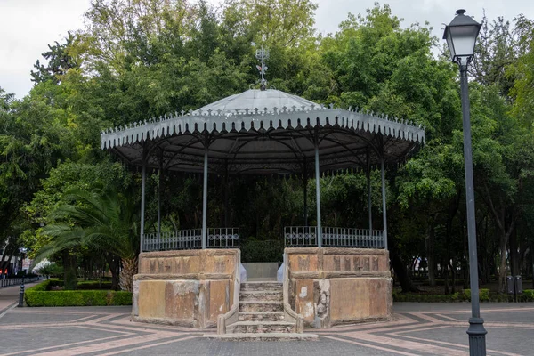 A small gazebo with a metal roof sits in a park. The gazebo is surrounded by trees. Historic center of Queretaro City, decorations and traditions to celebrate Mexico's Independence Day, colonial architecture, alleys and facades
