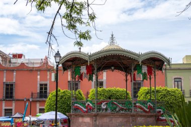 A small stage with a red roof and green plants. The stage is surrounded by a fence. Historic center of Queretaro City, decorations and traditions to celebrate Mexico's Independence Day, colonial architecture, alleys and facades