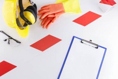 Red and white barrier tape strip separating construction safety gear including goggles, earmuffs, hard hat, and a pair of rubber gloves from a clipboard with blank sheet on white background clipart