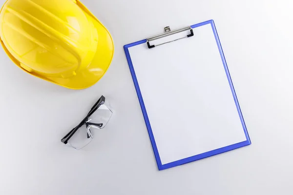 Hard hat, safety goggles, and a clipoboard with blank sheet flat lay on white background