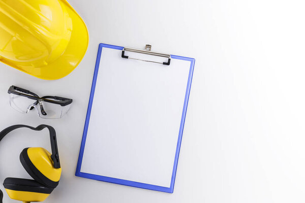 Clipboard with blank sheet alongside construction gear including hard hat, goggles, and earmuffs on a white background with copy space