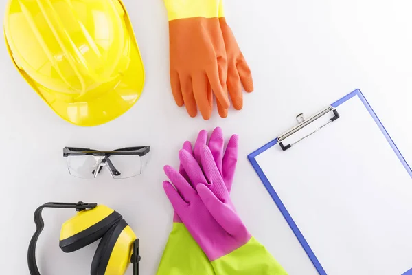 Hard hat, safety goggles, earmuffs, two pairs of rubber gloves, and a clipboard with blank sheet on white background