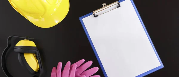 Banner image of clipboard with blank sheet of paper, hardhat, earmuffs, and gloves flat lay on black background