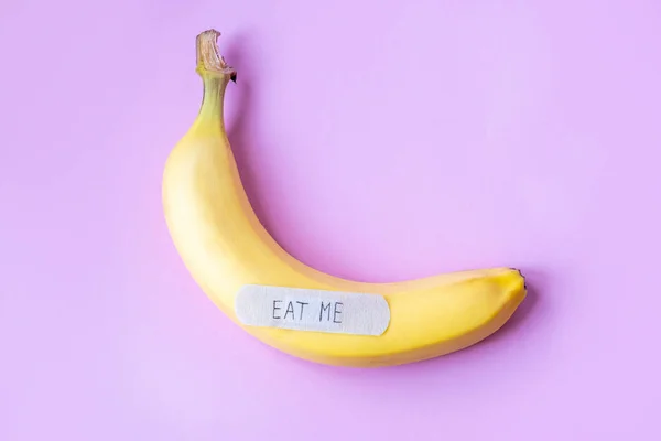 Yellow banana with plaster note EAT ME on pastel pink background. Minimal food concept. Flat lay, top view.