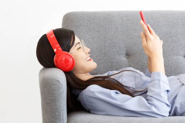 Neşeli Asyalı, kablosuz kulaklık takan akıllı telefondan müzik dinleyen neşeli kadının yan görüntüsü kanepeye uzandı..