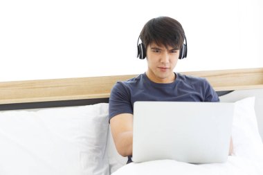 Mutlu Asyalı adam dizüstü bilgisayar kullanıyor ve yatak odasındaki iyi haberleri kontrol ediyor. Kopyalama uzayı. Takas. Film izliyordum. Ev kavramından çalış. Yeni Normal Kavram.