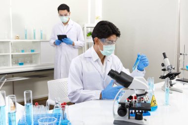 İki erkek bilim adamı yüz maskesi takıyor. Çözümü mikroskop ile kontrol ederken laboratuvarda çalışıyorlar. SARS-CoV-2, Covid-19 Teması.