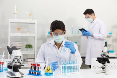 İki erkek bilim adamı yüz maskesi takıyor. Çözümü mikroskop ile kontrol ederken laboratuvarda çalışıyorlar. SARS-CoV-2, Covid-19 Teması.