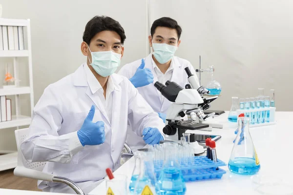Two Male Scientists wear Face Mask working in Lab while show thumb Up and Ready to Work and Presentation.  Blue Tone. SARS-CoV-2 , Covid-19 THEME.