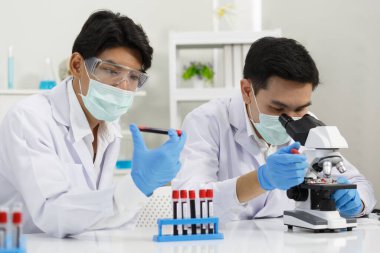 İki erkek bilim adamı, mikroskop tarafından test edilen kanı incelerken yüz maskesi takıyor. SARS-CoV-2, Covid-19 Teması.