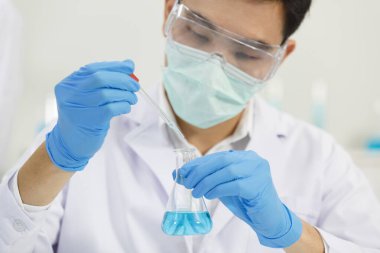 Erkek Asyalı Bilim Adamı, Erlenmeyer Matarası 'na Çözüm Bırakırken Laboratuar' da çalışırken maske takıyor. Mavi Ton. SARS-CoV-2, Covid-19 Teması.