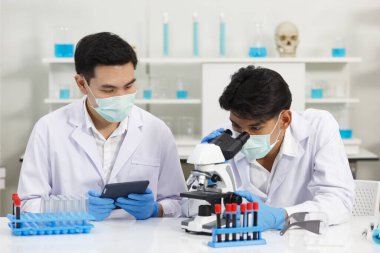 İki erkek bilim adamı yüz maskesi takıyor. Çözümü mikroskop ile kontrol ederken laboratuvarda çalışıyorlar. SARS-CoV-2, Covid-19 Teması.