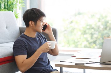 Dizüstü bilgisayarda çalışan mutlu Asyalı adam elinde bir fincan kahveyle evin zemininde oturuyor..